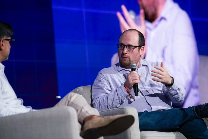 The image shows a man sitting in a chair with his arms outstretched while talking into a microphone. The background features a blue wall, which contrasts with the white shirt of another person who appears to be standing behind him. Both individuals are wearing glasses. There is also a text overlay on the image that reads "When the wallet disappears, everything else gets easier." This message seems to be related to the ongoing conversation between the two people in the scene.