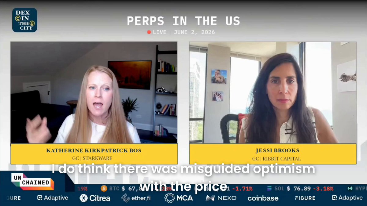 The image shows two women sitting across from each other, having a conversation or participating in an interview. They are positioned at opposite ends of a table with chairs placed around them. The background features a TV screen displaying the text "Perps In The US" along with some stock market data and a quote about Hyperliquid.