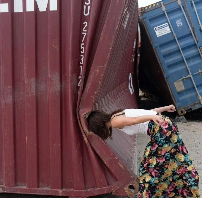 A woman in a white top and floral skirt bends backward dramatically between large red and blue shipping containers, with the number 'SU 27531 2' visible on the red container.