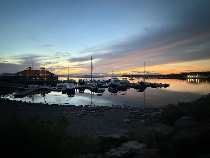 A serene marina at sunset with boats docked on calm water, with a building on the left and mountains in the distance.
