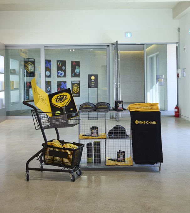 A BNB Chain pop-up display featuring branded yellow merchandise, hats, and swag in a shopping cart and on a wire rack set up in a building lobby. The "Wardrobe goals" caption highlights the trendy BNB Chain apparel and accessories as must-have fashion items.
