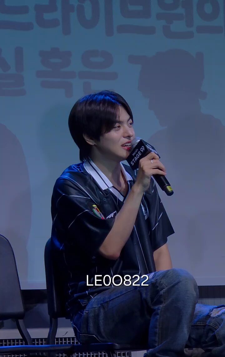 The image features a young man sitting down, wearing a black shirt with white stripes and blue jeans. He has his hand up to his mouth as if he's speaking into a microphone or giving a speech. The background of the photo shows a stage setting with a banner that reads "LEO at Melon STAGE 99". This suggests that the young man might be participating in an event, possibly related to the movie "Leo and the Lion", which is known for its Korean language elements.