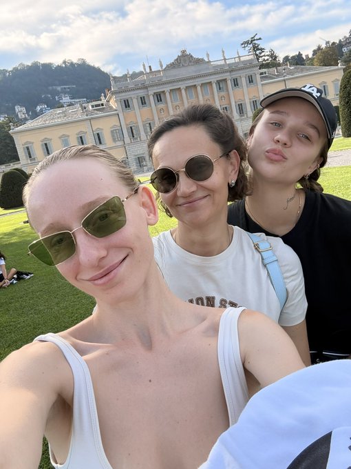 Selfie of three people posing outdoors in front of a grand yellow palace or villa with manicured gardens, mountains visible in the background.