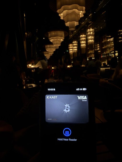 A smartphone displays a virtual Bitcoin debit card branded "KAST VISA INFINITE" held up to a card reader in a dimly lit, upscale bar, illustrating the post's message of spending Bitcoin in everyday luxury settings rather than selling it.