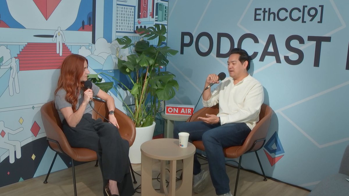 The image shows a man and woman sitting in chairs, talking to each other while holding microphones. They are positioned near a table that has a cup on it. The background features a wall with a poster for an EthcC podcast. The text "ethc(9) Podcast" is visible on the poster, indicating the topic of their conversation or discussion.