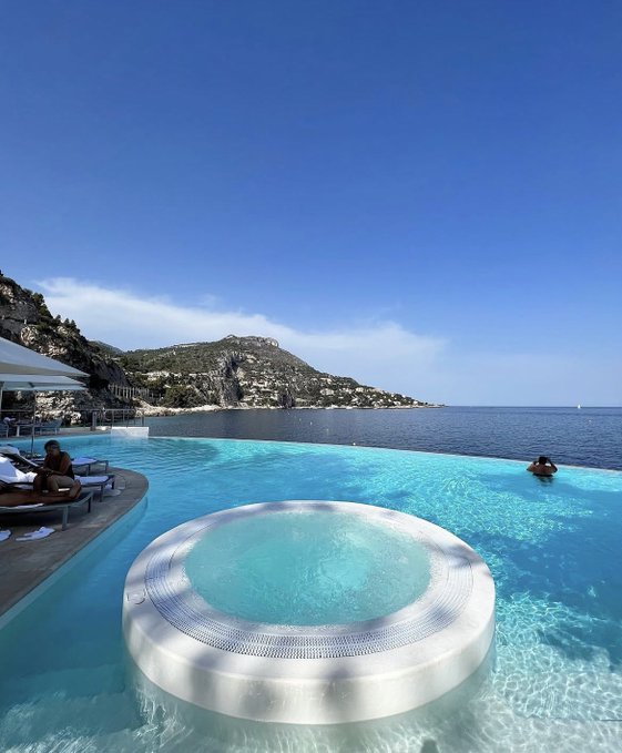The image shows a large swimming pool with a jacuzzi tub at its center. The pool is surrounded by chairs and umbrellas for relaxation. In the background, there are mountains visible against a clear blue sky. A person can be seen sitting in the water of the pool, enjoying their time.