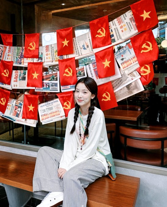 A young woman with braided hair sits on a wooden bench in front of a glass window decorated with Vietnamese flags (featuring yellow stars) and Communist Party flags (with hammer and sickle), alongside Vietnamese newspaper clippings. The flags suggest she may be celebrating Vietnam's National Day or a related patriotic holiday, fitting the cheerful "GM good morning" and holiday wishes in the post.