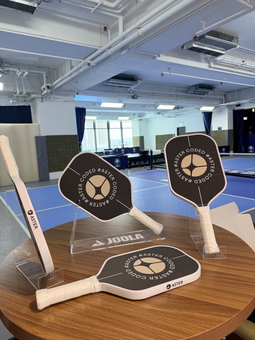 Three pickleball paddles branded 'ASTER #ASTER CODED' displayed on a round wooden table with a JOOLA logo, set in a sports facility.