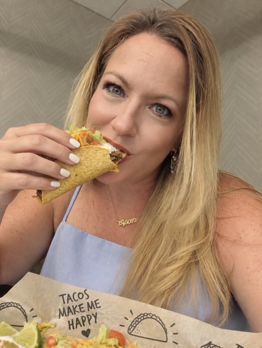 A blonde woman wearing a light blue top and a 'Bloom' necklace takes a bite of a taco, with the visible food wrapper reading 'TACOS MAKE ME HAPPY'.