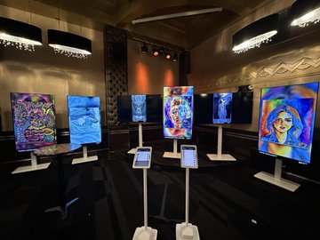 A dimly lit exhibition space with chandeliers, displaying multiple colorful digital artworks on white stands around a room, with an iPad on a stand in the foreground.