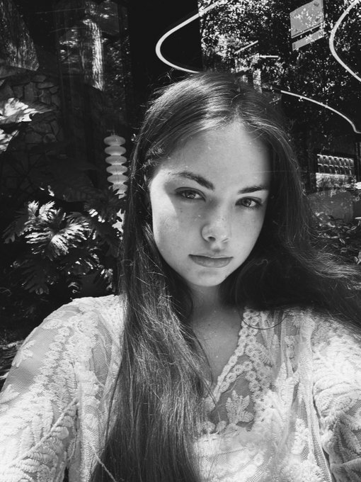 A black and white portrait of a young woman with long hair wearing a lace top, taken outdoors with foliage visible behind her.