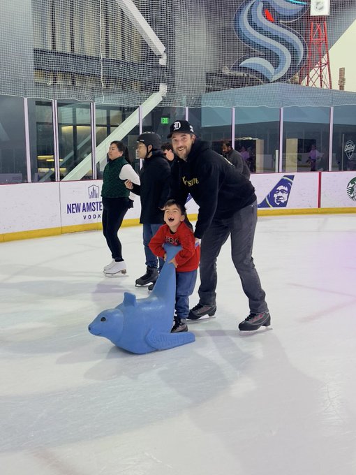 A smiling man in a black hoodie pushes a young boy in a red shirt who is riding a blue seal-shaped skating aid across the ice at an indoor skating rink, with 'NEW AMSTERDAM VODKA' visible on the boards.