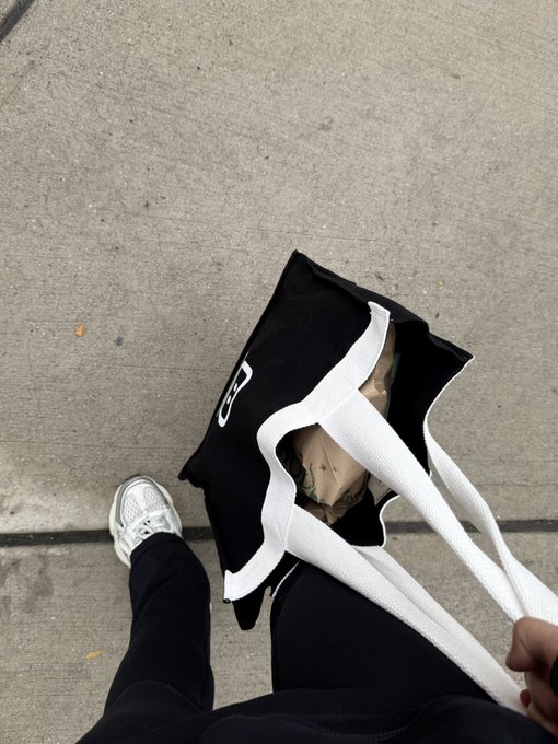 A person wearing white sneakers walks on concrete while carrying a torn black Nike tote bag with a broken handle.