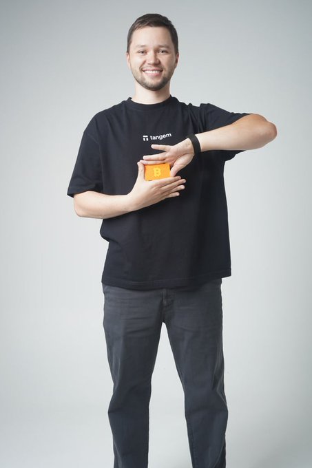 A smiling man wearing a black "Tangem" t-shirt holds up an orange Tangem crypto wallet in a celebratory pose, marking his 6-month work anniversary at the company.