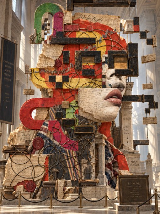 A complex mixed-media collage artwork displayed in a grand hall features a fragmented woman's face intertwined with colorful geometric shapes, architectural elements, and text fragments like 'We the People', 'MIKO LANIVII', and a 'RJ3RDR' plaque.