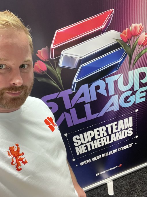 Man with blond hair and a beard wearing a white t-shirt with a red lion logo, taking a selfie in front of a 'Startup Village - Superteam Netherlands - Where web3 builders connect' poster featuring a Superteam logo and tulips.