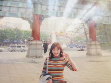 A blurry photograph of a smiling young woman in a striped sweater posing with a peace sign in front of a traditional gate with red pillars, with a van visible in the background.