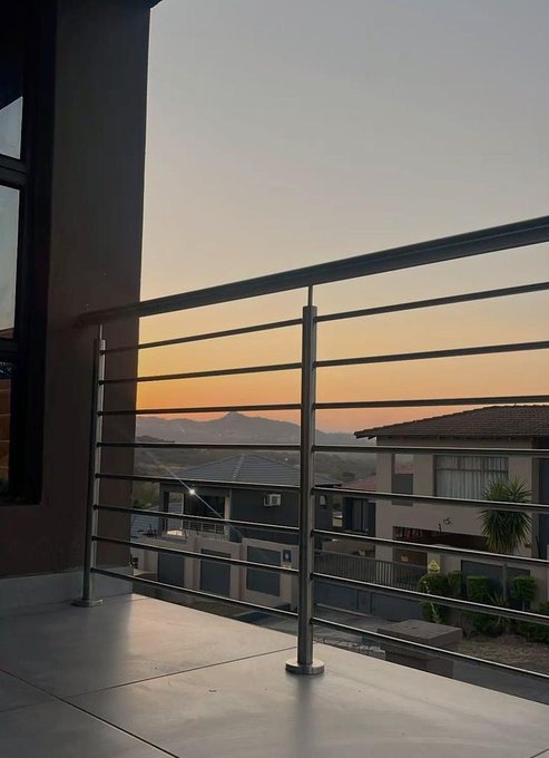 A balcony view at sunset or sunrise overlooking houses with a metal railing, featuring an orange and blue sky over distant mountains.