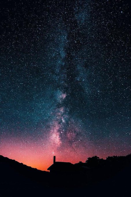 A stunning astrophotography shot captures the Milky Way galaxy stretching across a starry night sky, with a reddish glow near the horizon silhouetting a building and trees below.