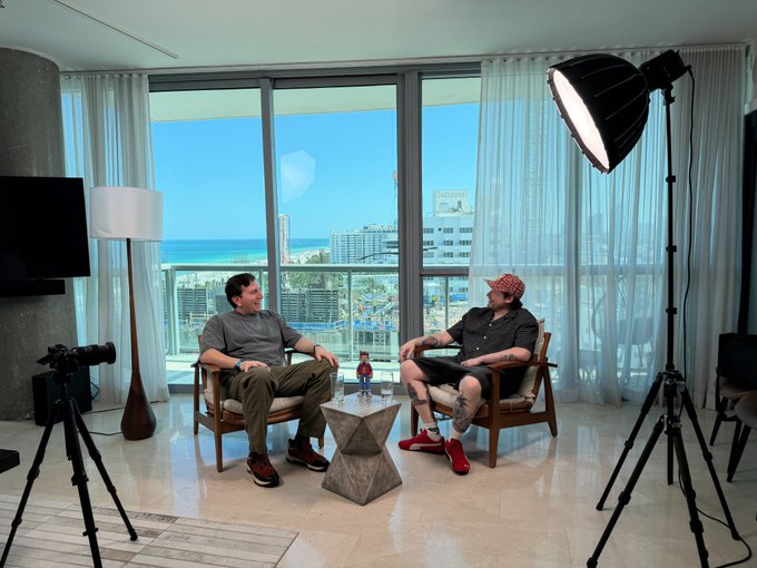 Two men sit in mid-century modern chairs having a conversation in a high-rise condo with a stunning ocean view, while a professional camera on a tripod and a large softbox light are set up in the foreground to film them.