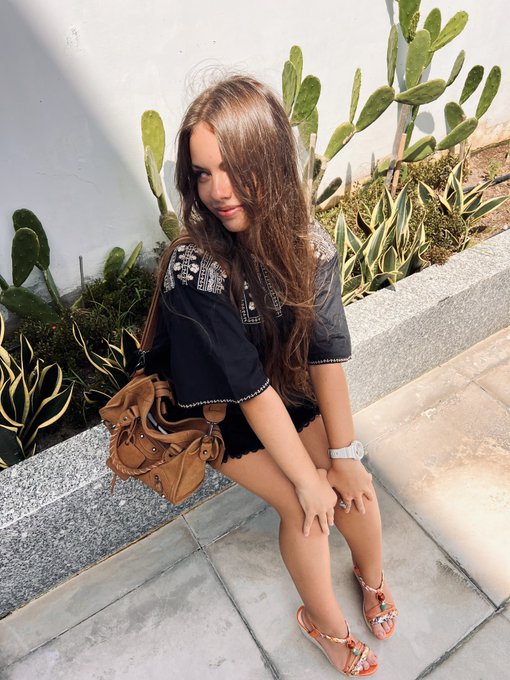 Anastasia poses in a dark embroidered blouse and floral sandals while leaning against a planter of cacti, her flowing long hair and relaxed pose echoing the artistic, laid-back vibe of a young creative. Her casual styling and natural setting align well with her intro as someone who enjoys drawing.