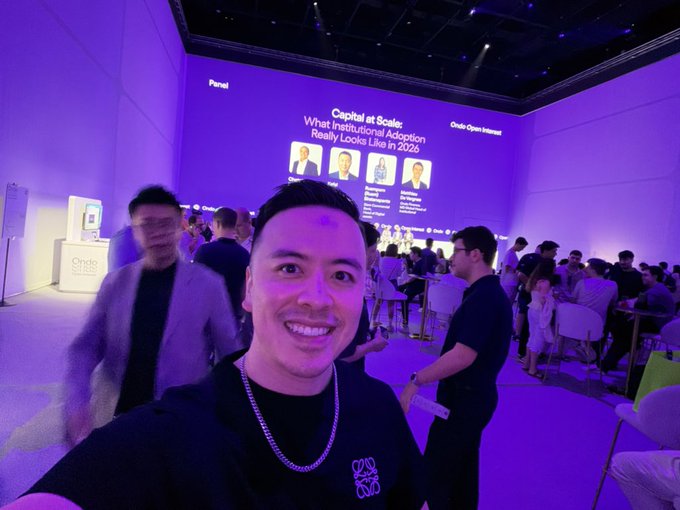 A man poses for a selfie at the Ondo Open Interest event during Token2049, with a large screen behind him displaying the panel discussion "Capital at Scale: What Institutional Adoption Really Looks Like in 2026," capturing the onchain trading competition atmosphere he references in his caption.