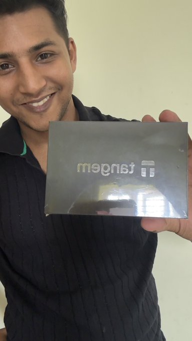 The image features a man holding up a small black box, which appears to be an electronic device or gadget. The man has a smile on his face as he poses with the item, possibly excited about its release or features. The background of the photo is white, and there are no other objects in the scene.