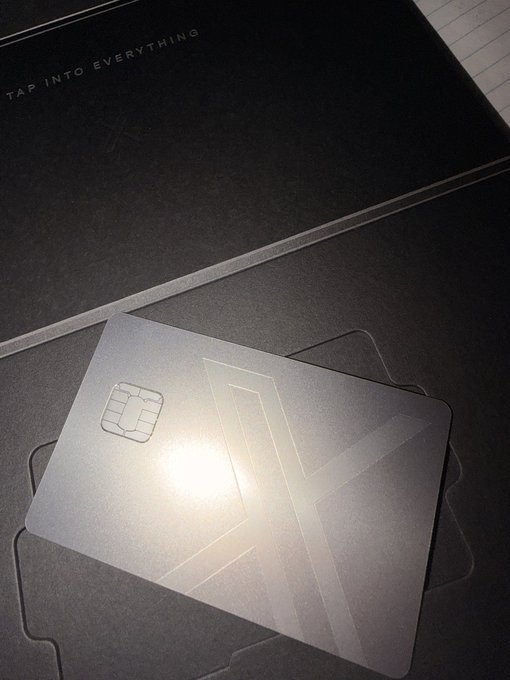 A photograph of a sleek dark metal card with an embedded smart chip and subtle etched X-design, resting on a dark textured surface near packaging that reads 'TAP INTO EVERYTHING.'