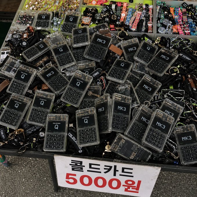 A pile of "MK3" and "COLDCARD Q" keychains, reminiscent of old calculators, are displayed for sale at Dongmyo for 5000 won. The image shows a close-up of these items, highlighting the nostalgic appeal that will soon be seen at Dongmyo.