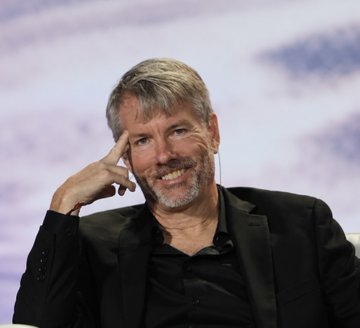 A man with gray hair and a beard, wearing a black blazer, sits thoughtfully with his hand on his temple against a cloudy backdrop. The image accompanies breaking news about Strategy (MSTR), likely featuring its co-founder Michael Saylor, announcing the company's purchase of 950 BTC worth $75 million, bringing its total holdings to 846,000 BTC valued at $71.9 billion.