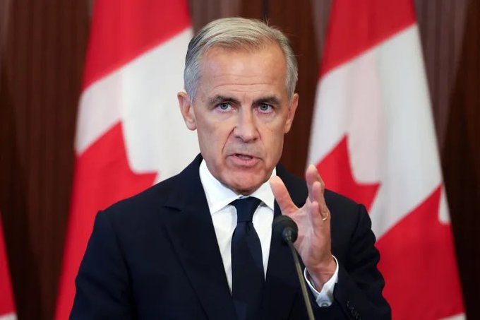 A man in a dark suit and tie, resembling Canadian politician Mark Carney, speaks at a podium with Canadian flags in the background.