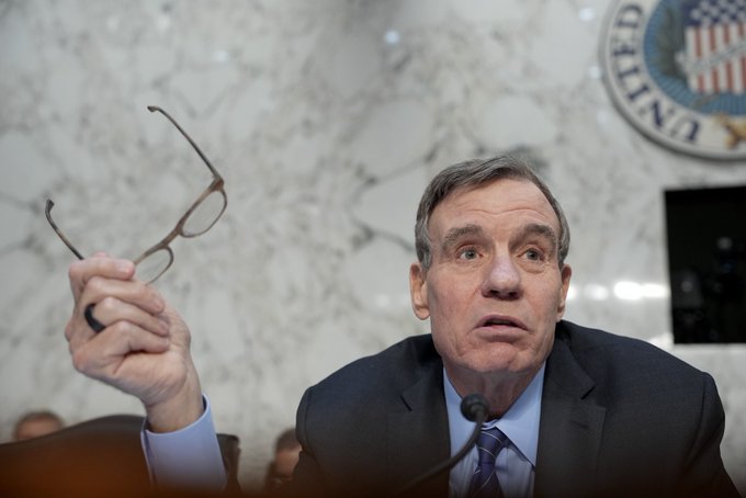 A man in a suit holds up a pair of glasses while speaking at what appears to be a United Nations hearing, with a US seal visible on the marble wall behind him.