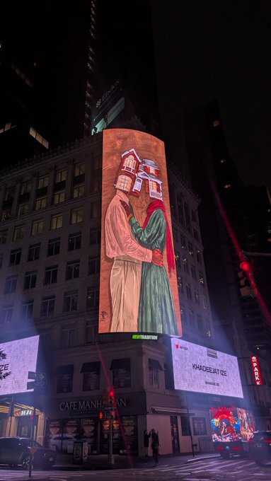 A nighttime street photo of a large illuminated billboard on a building displaying an artistic illustration of two robed figures, with "KIA" and "YAHOO!" signs visible.