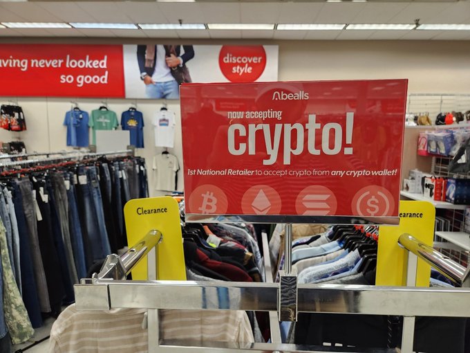 A 'now accepting crypto!' promotional sign at Bealls department store, declaring it the '1st National Retailer to accept crypto from any crypto wallet!' displayed near a clearance clothing rack.