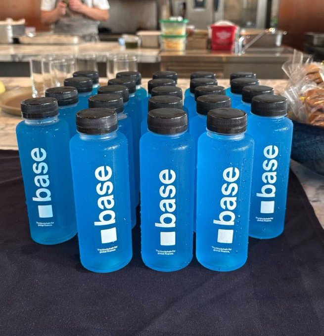Multiple blue "base" branded water bottles are arranged in rows on a black table in what appears to be a commercial kitchen or hospitality area.