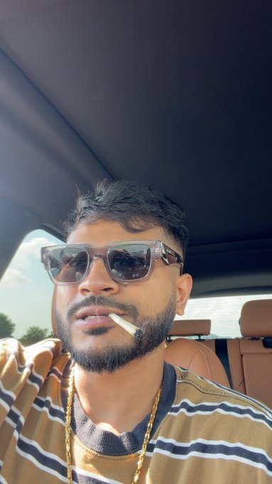 A man with a beard and sunglasses sits in a car, smoking a cigarette and wearing a striped shirt with a gold chain.