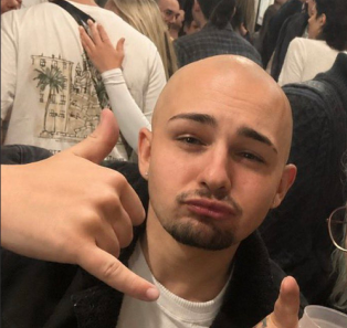 A selfie of a bald man with a beard making a 'call me' hand gesture with both hands, surrounded by other people in a crowded indoor setting.