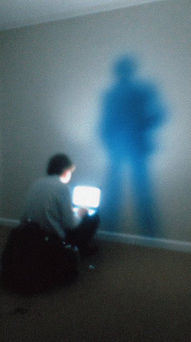 A blurry, grainy photograph of a person sitting on the floor using a laptop, with their large blue shadow cast on the wall behind them.