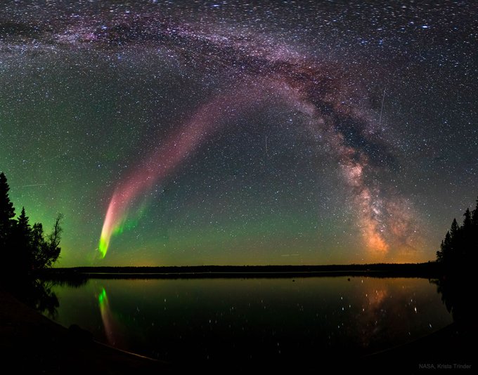 A stunning night sky over a lake showing the Milky Way galaxy and a green aurora, with credit text 'NASA, Krista Trinder' in the bottom right corner.