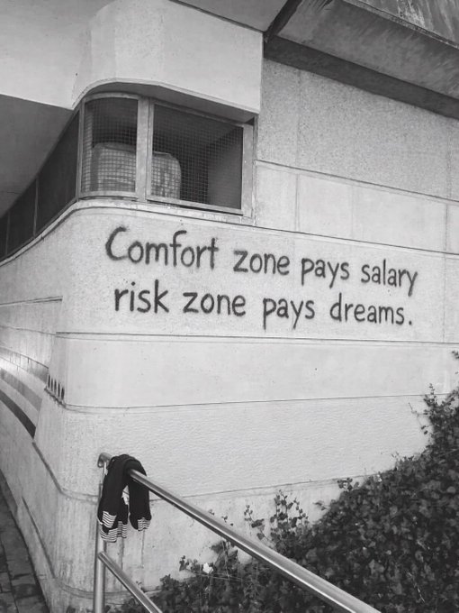 A black and white photo of a building wall with graffiti reading 'Comfort zone pays salary, risk zone pays dreams.' with a jacket hanging on a railing below.