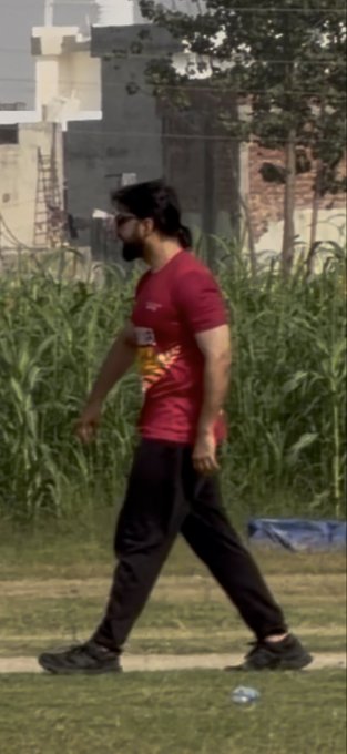 A blurry, cropped video still of a person with a beard walking outdoors past a cornfield and city wall, dressed in a red shirt and black pants.