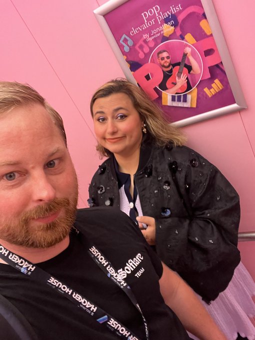 The image features a man wearing a black shirt and a woman wearing a black jacket standing next to each other, both smiling for the camera. They are positioned near an elevator with a poster of a man playing guitar hanging above them. The caption on the image reads "GM Almost weekend, Make sure you have plans with your loved ones (I'M gonna spend time in my @solflare wallet)".