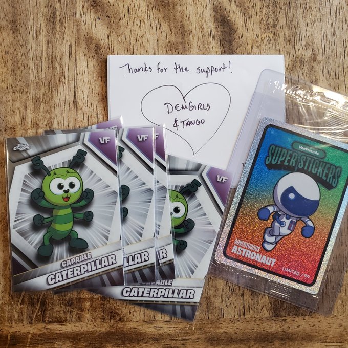 A handwritten thank you note addressed to 'Demigirls & Tango' is placed on a wooden surface alongside trading cards featuring a 'Capable Caterpillar' character and a holographic 'Adventurous Astronaut' sticker.