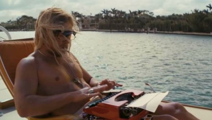 A vintage-style photograph shows a woman with long blonde hair wearing sunglasses while typing on an orange typewriter, seated on a boat with a waterfront and tropical scenery in the background.