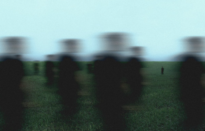 A motion-blurred photograph showing dark silhouettes of people sitting in a grassy field under a pale blue sky.