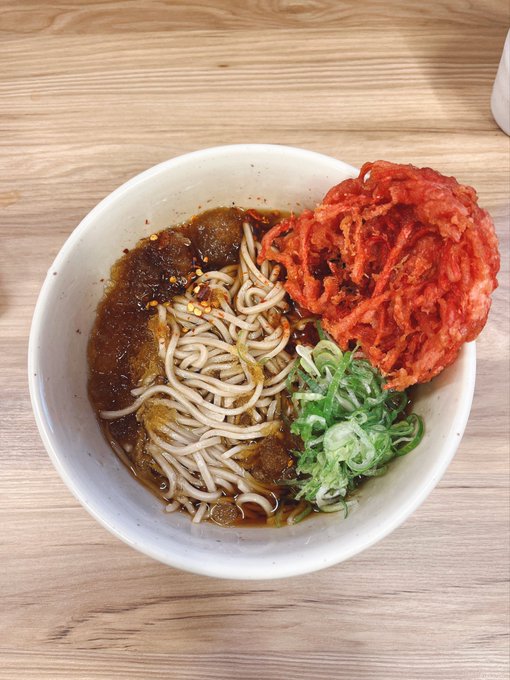 Overhead photo of a white bowl containing noodles in brown broth, topped with fried kimchi and chopped green onions on a wooden table.