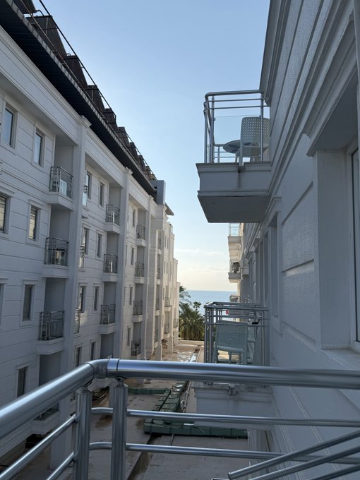A view from a balcony between two modern white apartment buildings, showing metal railings, air conditioning units, and a glimpse of the ocean in the distance under a clear sky.