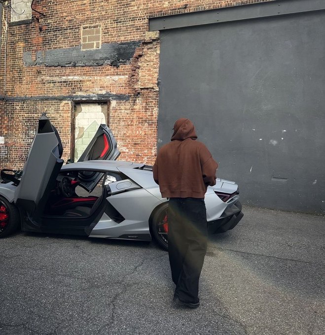 A person in a brown hoodie and dark pants stands next to a silver Lamborghini with its scissor doors open, parked against a brick wall.