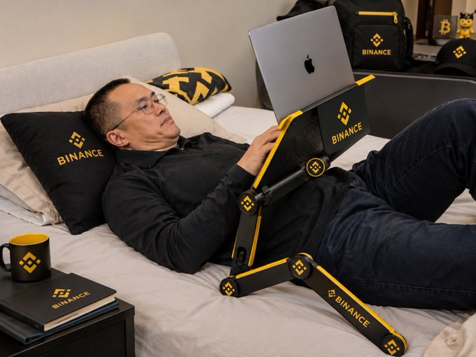 A man with glasses reclines on a bed using a laptop on a portable stand, surrounded by Binance-branded merchandise including pillows, a mug, and a notebook.