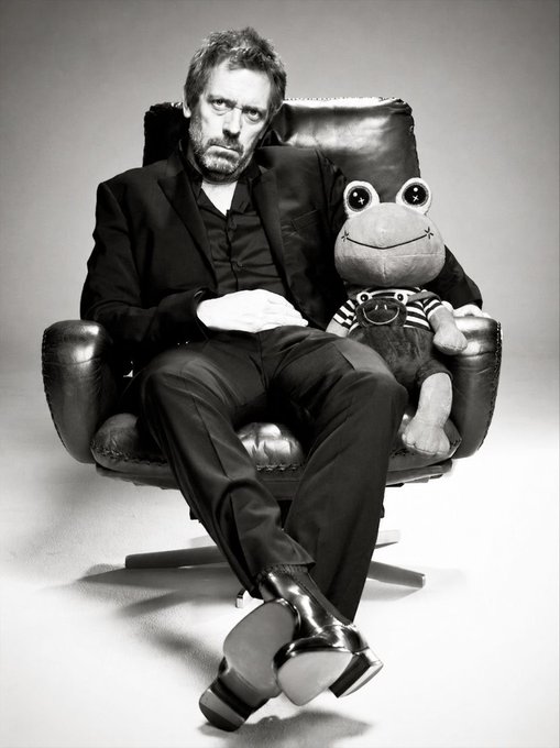 A black-and-white photograph of actor Hugh Laurie sitting in a leather chair with a large stuffed frog wearing goggles, both dressed in suits.